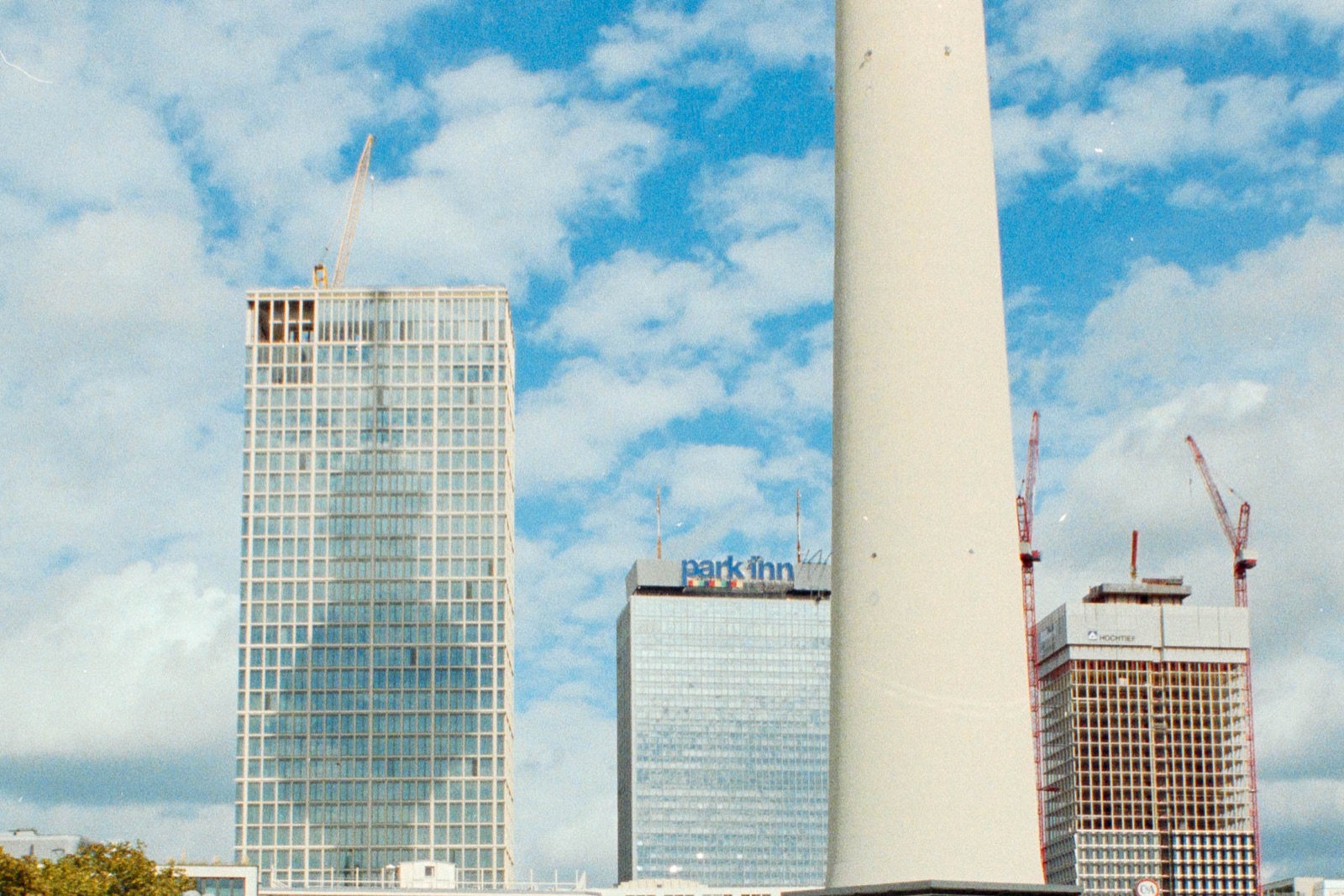 Berliner Fernsehturm im Anschnitt vor Hochhäusern mit Baukränen, darunter das Park Inn, blauer Himmel mit Wolken