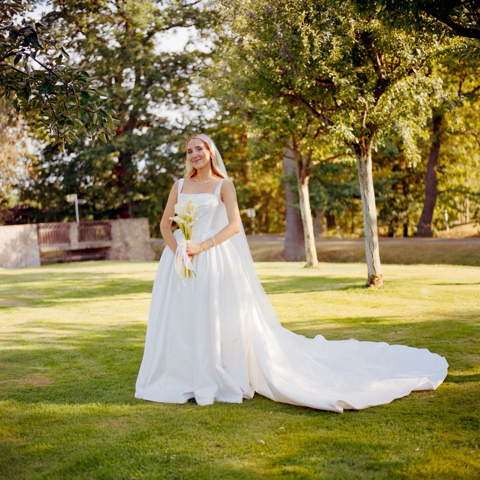 Braut im weißen Kleid mit langer Schleppe und Calla-Strauß auf einer sonnigen Wiese zwischen Bäumen