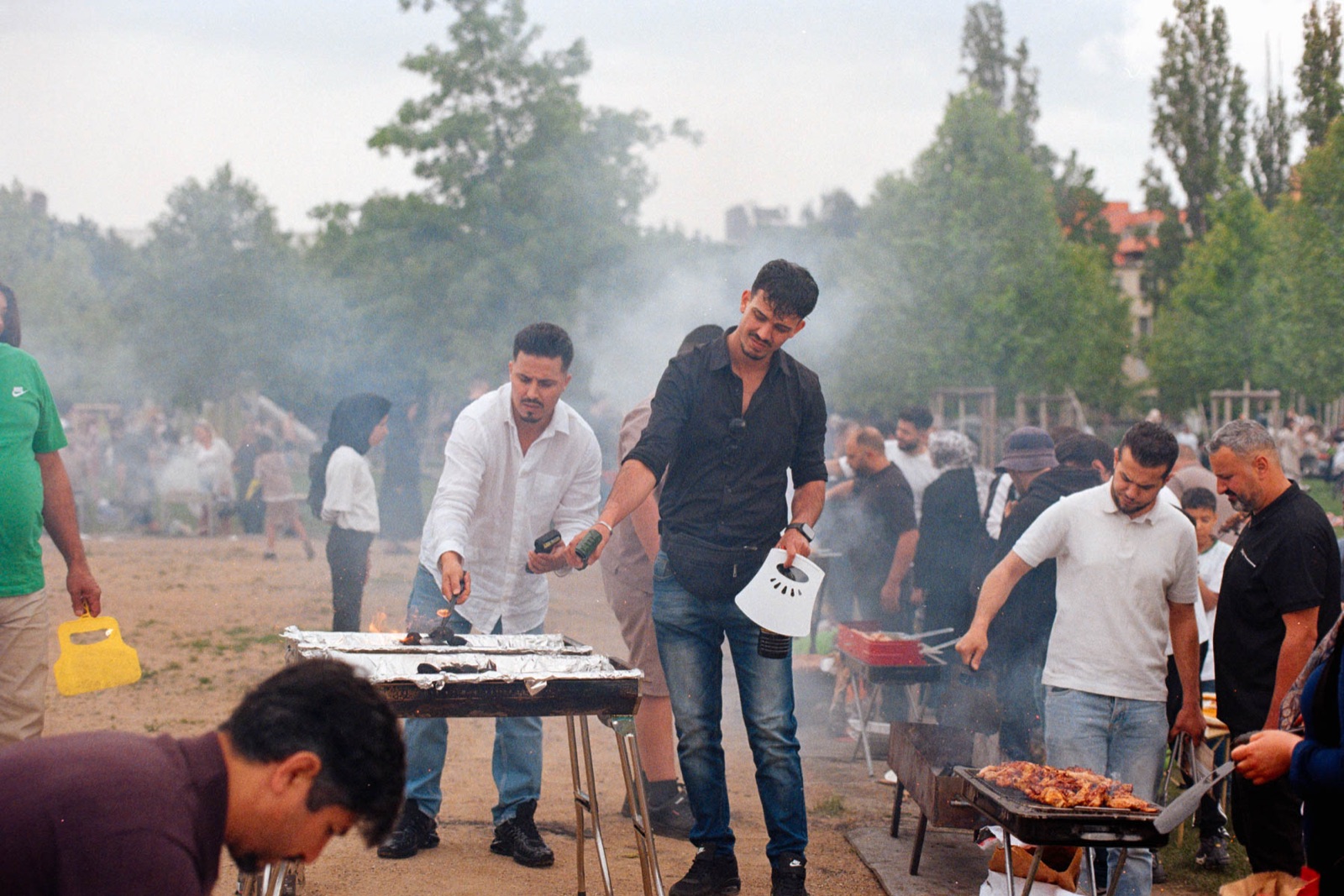 Männer grillen im Park, ein Mann fächert Rauch über einen Grill, dahinter viele Menschen und Bäume im Dunst