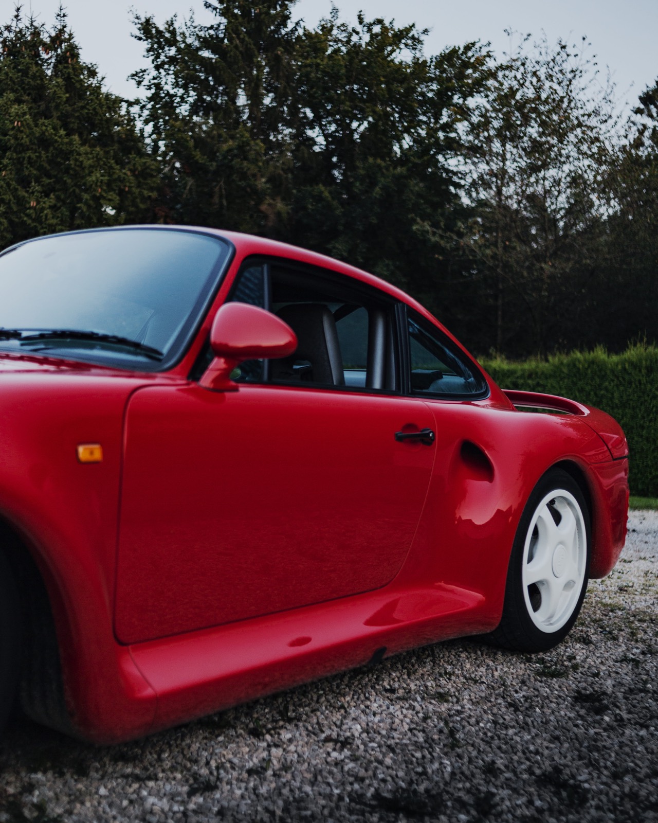 Roter Porsche 959 in der Seitenansicht auf einem Kiesplatz, im Hintergrund dunkle Tannen